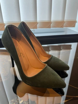 Mörkgröna pumps med spetsig tå - Snygga mörkgröna pumps med spetsig tå och hög klack. Skorna har en elegant siluett och är tillverkade i mockaimitation. Insidan är beige och de har en klassisk, stilren look som passar till många outfits.