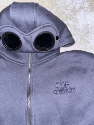 Blå hoodie med goggles från C.P. Company - Säljer en blå hoodie från C.P. Company med dragkedja och ikoniska goggles i huvan. Broderad logga på bröstet och mjukt material på insidan. Ena knappen på luvan gick av men jag satt dit den igen så det borde funka.  Storlek L men sitter som M.