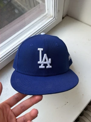 Blå LA Dodgers keps från New Era - Säljer en klassisk blå LA Dodgers keps från New Era med vit broderad LA-logga framtill och MLB-logga bak. Kepsen har rak skärm och är tillverkad i bomullstyg. Perfekt för dig som gillar streetwear och sportig stil. Borde kunna skicka samma dag men skriv vid minsta fundering! 