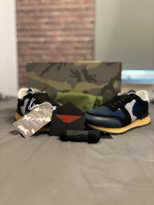 Valentino Garavani blå sneakers - Snygga sneakers från Valentino Garavani i blått, svart och vitt med camo-detaljer. Ovandelen är i mesh och mocka, med svarta skosnören och vit insida. Klassisk låg modell med beige sula och cool design som sticker ut.