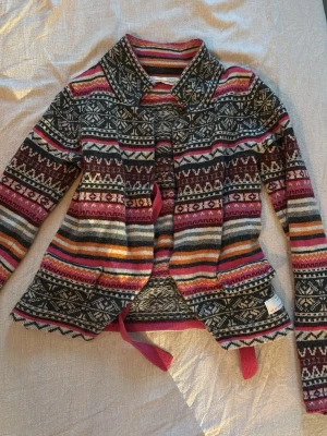 Mönstrad kofta med knytband - Färgglad kofta med traditionellt mönster i rosa, vinrött, orange, grått och vitt. Koftan har långa ärmar, krage och ett dekorativt knytband i midjan. Materialet ser ut att vara mjukt och stickat, perfekt för lager på lager.