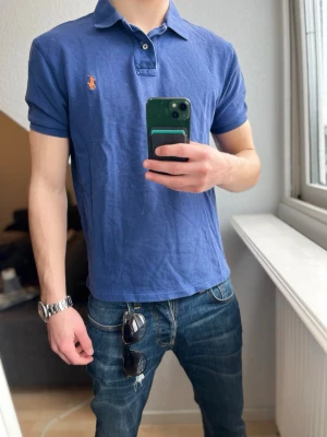 Blå pikétröja från Ralph Lauren - Klassisk blå pikétröja från Ralph Lauren med korta ärmar och den ikoniska broderade loggan på bröstet. Tröjan har krage och knappar vid halsen. Tillverkad i mjuk bomull som känns skön mot huden. Perfekt för en avslappnad och stilren look.