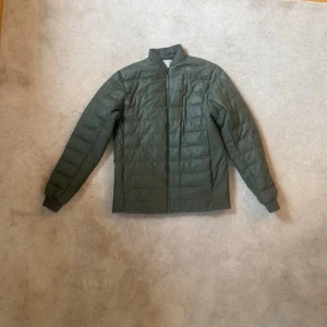 Rains Jacka - Snygg olivgrön pufferjacka med rak passform och diskret ståkrage. Jackan har quiltad design, hel dragkedja framtill och muddar vid ärmsluten. Perfekt för kalla dagar och enkel att matcha med olika outfits.