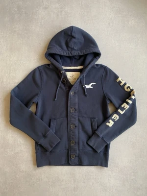 Mörkblå hoodie från Hollister - Mörkblå hoodie från Hollister med knappar framtill, två fickor och huva med dragsko. Stor logga i vitt på ena ärmen och Hollisters klassiska fågel på bröstet. Tillverkad i mjuk bomullsblandning för en skön och avslappnad look.