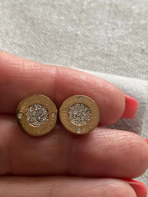 Guldtonade örhängen Michael Kors med Swarovski nya med box - Runda guldtonade örhängen från Michael Kors med gnistrande stenar i mitten och små stenar runt kanten. Märkets namn är ingraverat runt om. Snygg och stilren design som ger en lyxig känsla. Box och original intyg ingår.