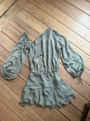 House of Cb klänning  - Grön klänning med oneshoulder-design och volangdetaljer. Klänningen har långa, puffiga ärmar med knyt vid handleden och en flowy kjol. Endast använd en gång 🫶