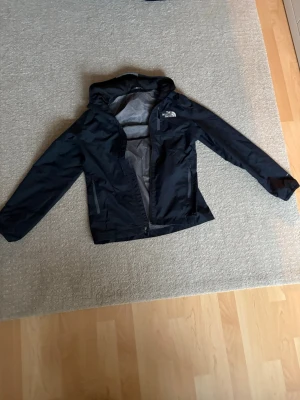 Svart vindjacka från The North Face - Snygg svart vindjacka från The North Face med huva och dragkedja framtill. Jackan har meshfoder, bröstficka med dragkedja och logga på bröstet. Perfekt för blåsiga dagar och enkel att packa ner i väskan.