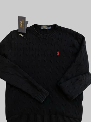 Svart kabelstickad tröja Polo Ralph Lauren - Kabelstickad svart tröja från Polo Ralph Lauren med rund halsringning och röd broderad logga på bröstet. Tröjan har ribbade muddar vid ärmslut och nederkant. Perfekt för en stilren och klassisk look.