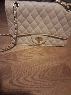 Beige quiltad handväska med kedja från Ur&Penn. - Snygg beige handväska i quiltat mönster med guldfärgad kedjerem och vridlås framtill. Klassisk design med elegant känsla, perfekt för dig som gillar stilrena accessoarer. 