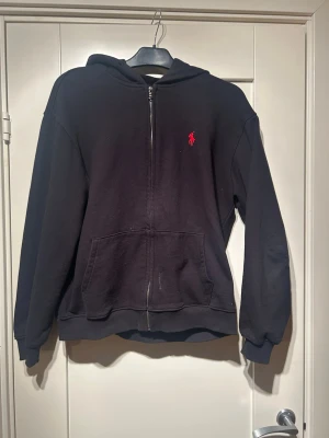 Svart hoodie från Polo Ralph Lauren - Klassisk svart hoodie med dragkedja från Polo Ralph Lauren. Modellen har huva, två fickor fram och det ikoniska röda Polo-märket broderat på bröstet. Tillverkad i mjukt bomullsmaterial! Den är storlek L men sitter som en m skulle jag säga.