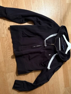Svart hoodie från Ralph Lauren - Svart hoodie med dragkedja från Ralph Lauren. Klassisk design med vit logga broderad på bröstet och vita snören i huvan. Mjuk bomullskänsla och två fickor framtill. Perfekt för en avslappnad och stilren look.