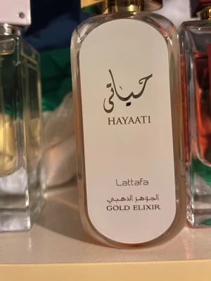 Lattfa Hayaati Gold Elixir parfym - Exklusiv parfym från Lattafa, Hayaati Gold Elixir, i en elegant rektangulär glasflaska med vit och guld detalj. Flaskan har en stilren design med guldfärgat lock och arabiska inskriptioner. Perfekt för dig som gillar lyxiga dofter.