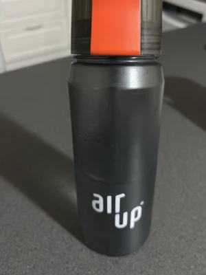 Svart Air Up vattenflaska - Säljer en svart Air Up vattenflaska med tydlig logga och orange detalj på locket. Flaskan är tillverkad i plast och har en modern, stilren design. Perfekt för att smaksätta vatten med doftpoddar. Locket är avtagbart och har en praktisk greppfunktion.