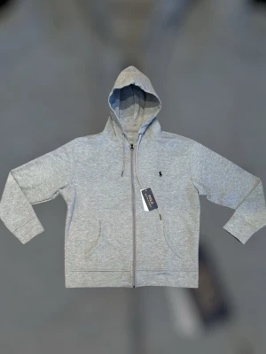 RL bundle - Snygg grå hoodie från Polo Ralph Lauren med dragkedja framtill, två fickor och justerbar huva med snören. Klassisk logga broderad på bröstet. Perfekt för en avslappnad och stilren look. Tillverkad i mjukt bomullsmaterial.