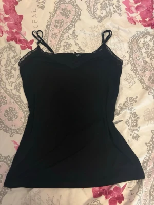 Svart spetsdetaljerad linnetopp - Säljer en svart linnetopp med tunna axelband och spetsdetaljer vid urringningen. Toppen har en figurnära passform och är perfekt för att styla lager på lager eller bära som den är. Enkel men ändå snygg design.