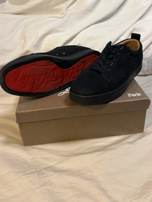 Svarta sneakers från Christian Louboutin - Snygga svarta sneakers från Christian Louboutin med klassisk röd sula och diskret logga. Skorna har lågt skaft, rund tå och är tillverkade i mocka med svarta skosnören. Perfekta för dig som vill ha en clean och exklusiv look. Storlek 42