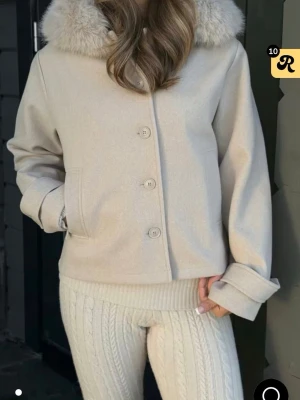 Beige ulljacka - Stilren beige jacka med stor, fluffig fuskpäls runt kragen och knäppning framtill. Jackan har raka ärmar, fickor på sidorna och en klassisk. Perfekt för kyliga dagar och enkel att matcha med olika outfits.