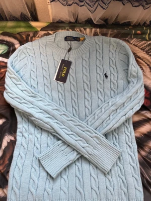 Ljusblå kabelstickad tröja Polo Ralph Lauren - Snygg ljusblå kabelstickad tröja från Polo Ralph Lauren med klassisk rund halsringning och broderad logga på bröstet. Tröjan har långa ärmar och ribbade muddar vid ärmslut och nederkant. Perfekt för dig som gillar stilren och tidlös design.