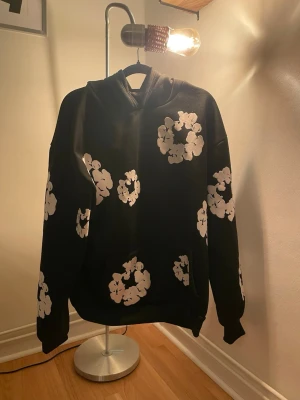 Demin tears hoodie  - Snygg svart hoodie med stora vita blommönster över hela plagget. Hoodien har en klassisk huva och ribbade muddar vid ärmar och nederkant. Perfekt för dig som vill sticka ut med en unik och trendig stil. Materialet ser ut att vara mjukt och bekvämt, troligen bomull eller bomullsblandning osäker på äktheten därav priset..