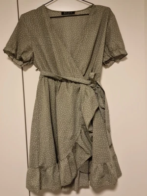 Grön prickig omlottklänning - Söt grön kortklänning med vita prickar och volangkant. Klänningen har omlottmodell med knyt i midjan och korta puffärmar. Perfekt för sommarens alla tillfällen och ger en fräsch och ungdomlig look.