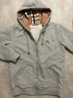 Grå Burberry zip up tröja - Snygg grå zip hoodie från Burberry med klassiskt rutigt foder i huvan och broderad logga på bröstet. Hoodien har dragsko i huvan, två stora fickor fram och ribbade muddar. Perfekt för dig som gillar stilrena och exklusiva plagg.