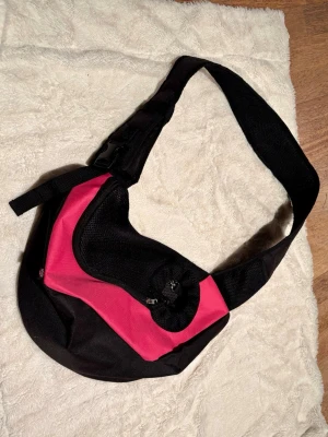 Svart och rosa axelväska för en liten hund eller en katt - Cool axelväska i svart och rosa med justerbar rem och praktiskt spänne. Väskan har dragkedja och snörning upptill samt en sportig, rundad form. Tillverkad i slitstark textil, perfekt för dig som vill ha en färgklick till outfiten.