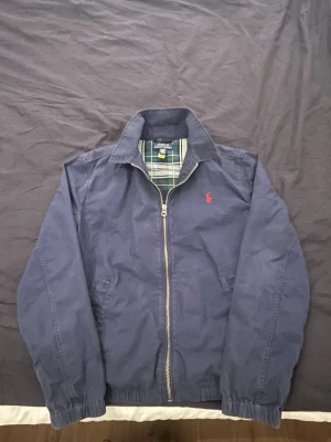 Marinblå vindjacka från Polo Ralph Lauren - Snygg marinblå vindjacka från Polo Ralph Lauren med dragkedja, klassisk krage och röd broderad logga på bröstet. Jackan har rutigt foder i grönt och vitt samt elastiska muddar vid ärmslut och midja. Perfekt för en stilren och avslappnad look.