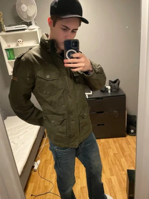 Olivgrön vindjacka från Crafted Goods - Snygg olivgrön vindjacka från Crafted Goods med fyra framfickor med lock och knapp, hög krage och dragkedja. Jackan har en klassisk militärinspirerad look och är tillverkad i ett slitstarkt material med svart innerfoder. Jackan är för stor och jag är ca 186 70 kg✌🏼