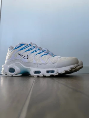 Nike Air Max Plus vita/blå sneakers - Nike Air Max Plus sneakers i vitt mesh och syntet med blå detaljer och svarta Swoosh-loggor på sidorna. Skorna har synliga Air-enheter i sulan och en sportig, futuristisk design. Perfekta för dig som gillar streetwear och vill sticka ut med en klassisk modell.