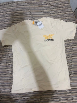 Gallery Dept t shirt - Säljer en beige t-shirt från Gallery Dept med tryck i gult och svart på bröstet. T-shirten har rund hals och korta ärmar. Tillverkad i mjuk bomull som känns skön mot huden. Perfekt för dig som gillar streetwear och enkel stil.