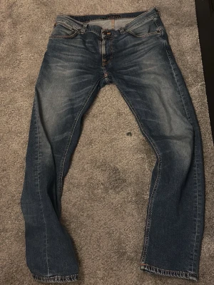 Blå jeans från Nudie Jeans - Säljer ett par klassiska blå jeans från Nudie Jeans med snygga slitningar och kontrastsömmar. Jeansen har rak passform, fem fickor och detaljerad bakficka med Nudie-logga. Perfekta för en avslappnad och trendig stil.