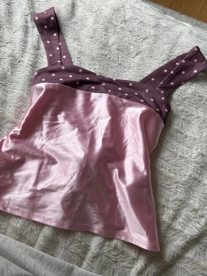 Rosa satintopp med prickiga band - Söt linnetopp i glansigt rosa satin med breda axelband och kant i mörklila tyg med vita prickar. Toppen har knytband i ryggen för justerbar passform och en feminin look. Perfekt för dig som gillar unika detaljer.