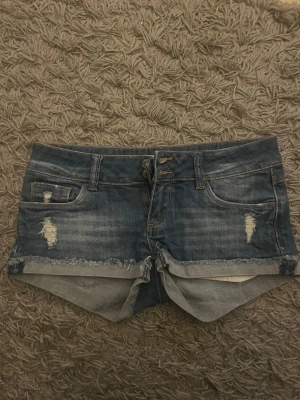 Lågmidjade shorts - Säljer ett par blå lågmidjade jeansshorts med slitna detaljer och råa kanter. Shortsen har klassisk femficksdesign, knappgylf och snörning framtill. Passar inte mig då jag köpte dessa second hand så har tyvärr inga bilder med shortsen på. Skicka gärna om ni vill ha mer info eller flera bilder!💌