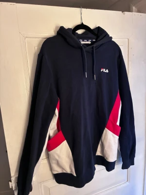 Mörkblå hoodie från FILA med färgblock - Snygg mörkblå hoodie från FILA med huva och dragsnören. Tröjan har vita och röda färgblock på sidorna med öppna fickor och en liten FILA-logga broderad på bröstet. Tillverkad i mjukt bomullsmaterial, perfekt för en avslappnad och sportig stil.