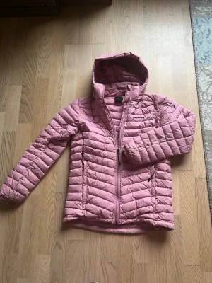 Rosa dunjacka från Peak Performance - Säljer en rosa dunjacka från Peak Performance med huva och dragkedja framtill. Jackan har två sidofickor med dragkedja och quiltad design. Perfekt för kalla vinterdagar och riktigt snygg färg som sticker ut.