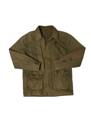 Fred Perry overshirt - Det finns red på insidan som man kan göra den tajtare runt midjan Stilren olivgrön overshirt i bomull med klassisk krage och två stora fickor framtill. Jackan har knappstängning vid ärmsluten och en avslappnad passform. Perfekt lager på lager-plagg för en cool och enkel look.