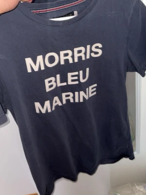 Morris Bleu Marine t-shirt marinblå - Snygg marinblå t-shirt från Morris med vit text 'MORRIS BLEU MARINE' på bröstet. Klassisk rund halsringning och korta ärmar. Perfekt för en avslappnad och stilren look. Inga tecken på användning. Hör av dig om du har några frågor.