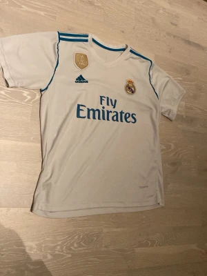 Real Madrid matchtröja Adidas vit - Säljer en vit Real Madrid matchtröja från Adidas med blå detaljer och FIFA World Champions 2017-märke på bröstet. Tröjan har klubbmärke, Adidas-logga och Fly Emirates-tryck på framsidan. Skickar inom 24 Tim!😊💨