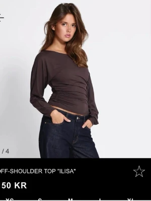 Mörkbrun offshoulder topp  - Trendig mörkbrun offshoulder topp 'Ilisa' med lång ärm och rynkade detaljer i midjan. Sitter tight över kroppen och har en snygg asymmetrisk axel. Perfekt att styla med jeans eller kjol för en cool look.
