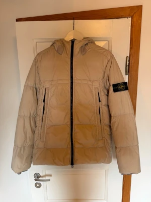 Beige pufferjacka från Stone Island - Säljer en beige pufferjacka från Stone Island med huva och svart logotyp-patch på vänster ärm. Jackan har två stora framfickor med dragkedja och en rak passform. Materialet är quiltat och vadderat för extra värme, perfekt för kalla dagar. Inköpt på NK Stockholm 2021 Nypris runt 11 000. Kvitto finns inte men QR kod på insidan av jackan för Authentic check. 