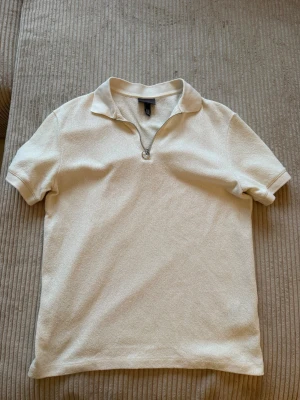 Beige kortärmad topp från H&M - Snygg beige topp från H&M med korta ärmar och krage. Toppen har en halv dragkedja framtill som ger en sportig touch. Tillverkad i ett mjukt, strukturerat material som känns skönt mot huden. Perfekt för dig som gillar enkel men stilren design.