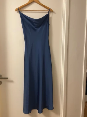 Blå långklänning med smala axelband - En stilren blå långklänning med smala axelband och mjukt fall. Klänningen har en enkel, elegant siluett och lätt glansigt material som ger en lyxig känsla. Perfekt för dig som gillar minimalistisk och tidlös stil.