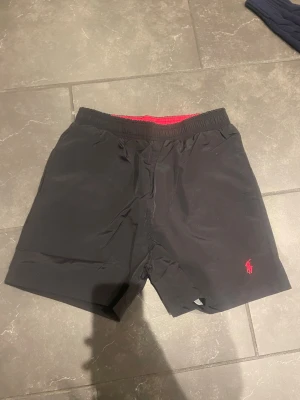 Svarta shorts från Ralph Lauren - Svarta shorts från Ralph Lauren med elastisk midja och röd logga broderad på vänster ben. Shortsen har en enkel och klassisk design med fickor på sidorna och röd insida i midjan. Perfekta för en avslappnad stil.