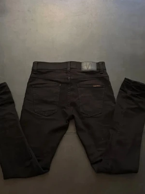 Nudie Jeans svarta - Säljer ett par svarta skinny jeans från Nudie Jeans med klassisk femficksmodell och diskret logga på bakfickan. Storlek: W29, L32 Jeansen har en smal passform och är tillverkade i stretchigt material för extra komfort. Hör av dig vid frågor elr liknande!