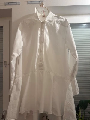 Vit skjortklänning med volang - Säljer en vit skjortklänning med lång ärm och volang nedtill. Klänningen har klassisk krage, knäppning framtill och är tillverkad i ett bomullsliknande material. Perfekt för dig som gillar en clean och enkel stil.
