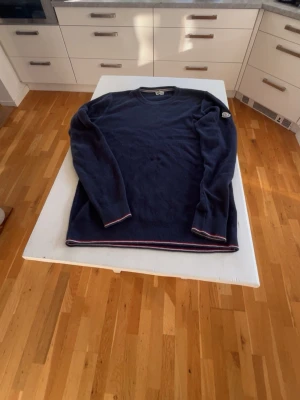 Mörkblå Moncler tröja  - Storlek S men egentligen M. Mörkblå stickad tröja från Moncler med rund halsringning och ribbade muddar. Tröjan har en ikonisk Moncler-patch på ärmen och diskreta ränder i rött och vitt vid hals och nederkant. Perfekt för dig som gillar stilrena och exklusiva plagg.