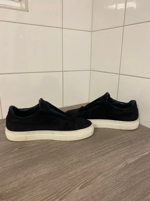 Mörkblå sneakers från Axel Arigato - Stilrena mörkblå sneakers från Axel Arigato i mocka med vit, chunky sula. Diskret logga på sidan och minimalistisk design utan synliga snören. Perfekta för dig som gillar en clean och modern look.