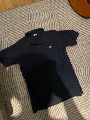 Mörkblå pikétröja från Lacoste - Klassisk mörkblå pikétröja från Lacoste med den ikoniska gröna krokodilloggan på bröstet. Tröjan har korta ärmar, krage och knäppning framtill. Perfekt för en stilren och avslappnad look.