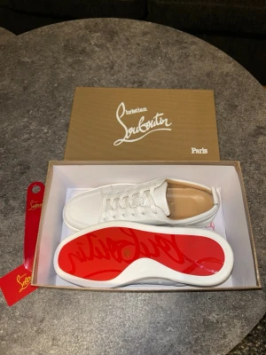 Vita sneakers från Christian Louboutin - Stilrena vita sneakers från Christian Louboutin med klassisk röd sula och diskret logga undertill. Skorna är låga och tillverkade i strukturerat läder med vita snören och detaljer. Modellnamn: Rantulow. Perfekta för dig som vill ha en clean look med exklusiv känsla. Helt nya. Priset är ej hugget i sten! VÄLDIGT SÄLLSYNTA PÅ PLICK