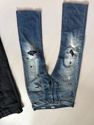 Blå slitna jeans från Replay - Säljer ett par blå jeans från Replay med kraftigt slitna detaljer och hål på benen för en riktigt edgy look. Jeansen har raka ben, råa kanter vid benslut och klassisk femficksmodell. Perfekta för dig som gillar streetwear och avslappnad stil.